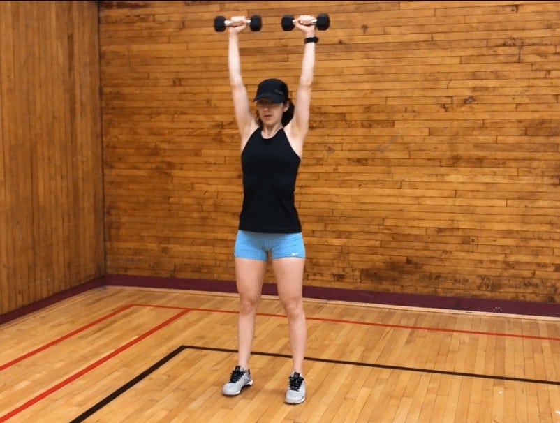 brittany pressing two dumbbells directly over her head as she stands tall