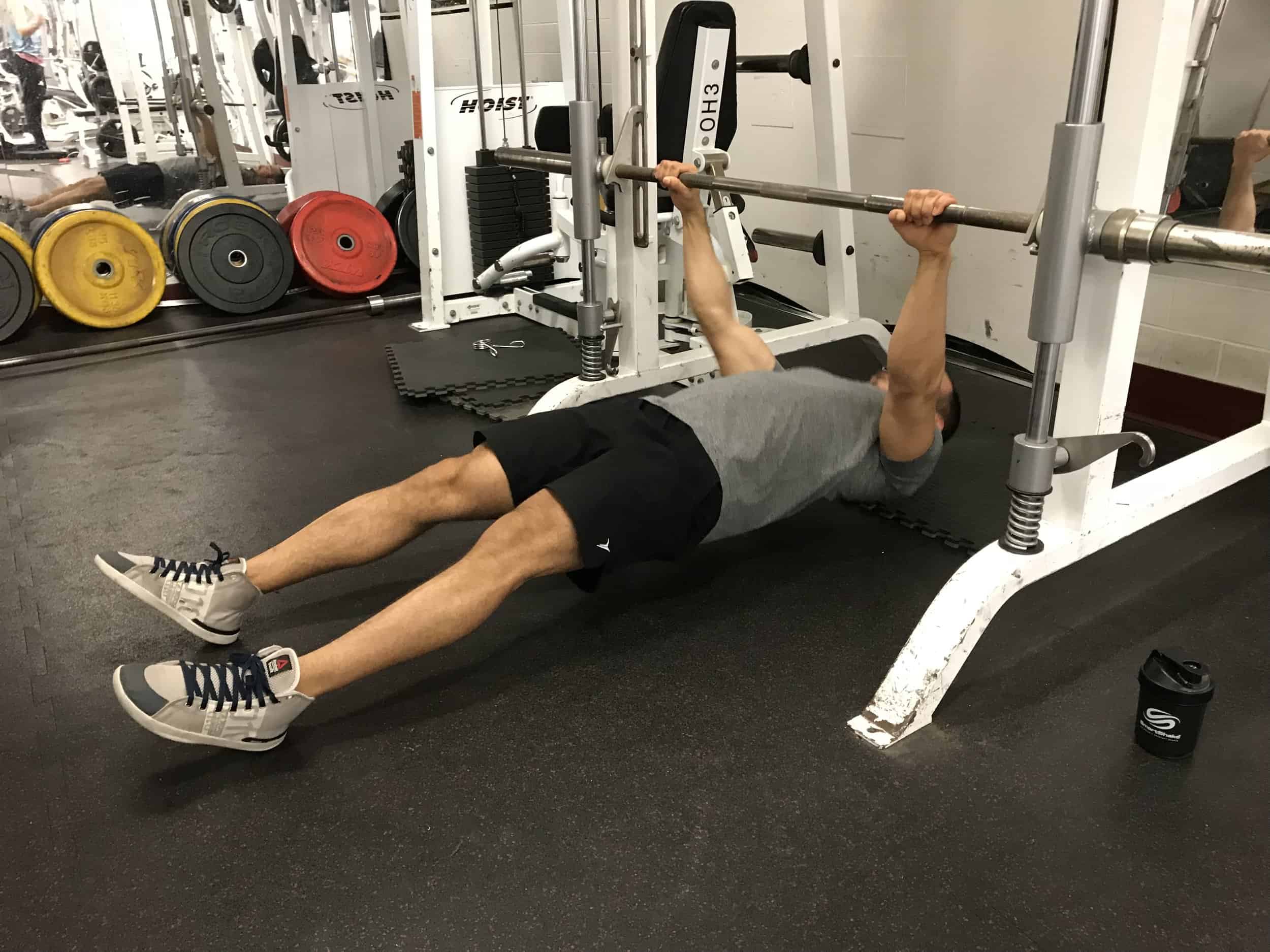 alex at the bottom part of a horizontal pullup hanging from a barbell with his arms straight and back parallel to the ground and feet straight