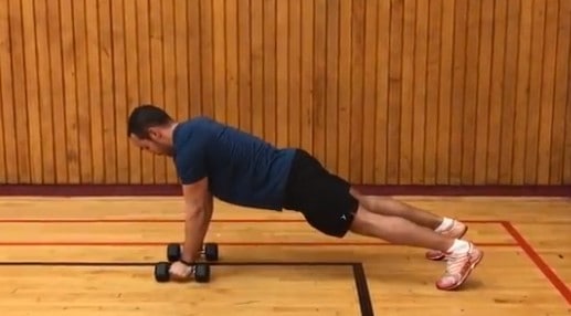 alex in a pushup position holding onto two dumbbells that are positioned on the floor underneath his hands