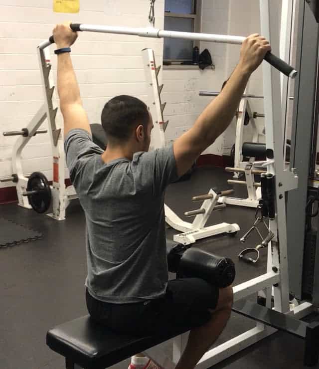 alex at the top of a lat pulldown with his arms extended overhead grabbing the bar and knees anchored under the pads