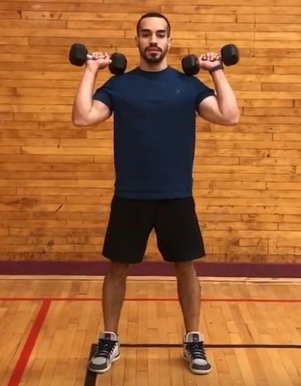alex standing tall holding a dumbbell on each hand at his shoulders with a stance slightly wider than shoulder-width