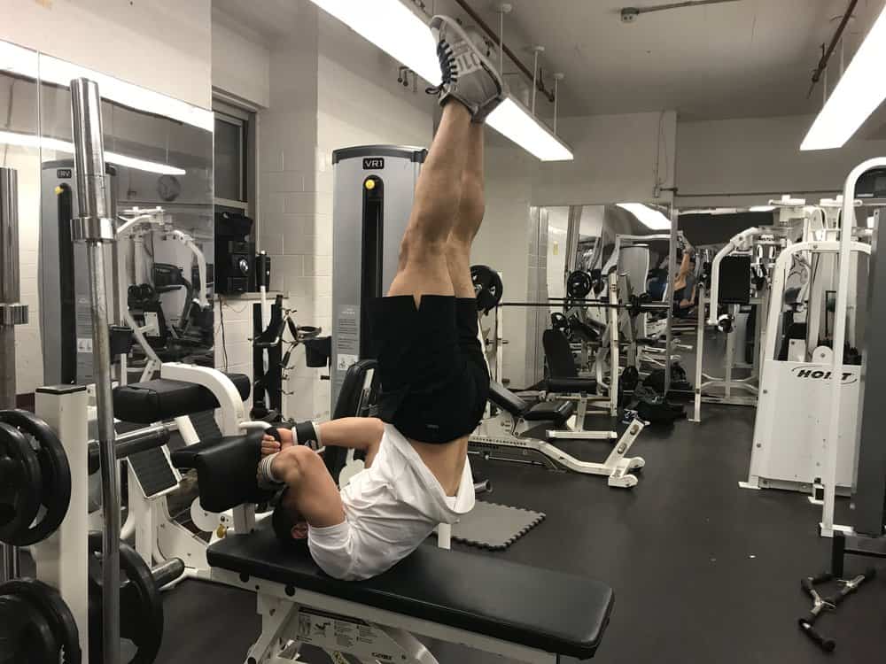 alex on his back holding onto something behind him while lifting his legs straight up to the sky without bending at the hips