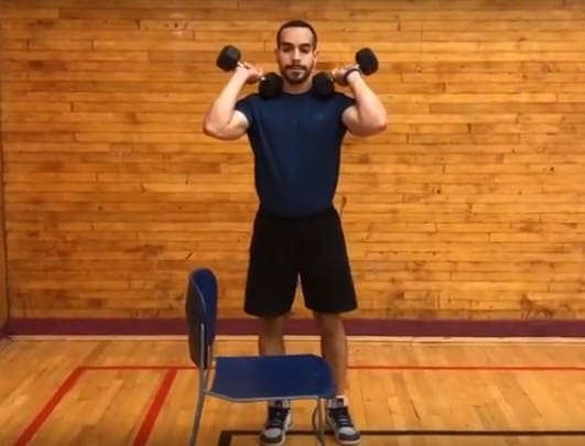 alex in front of a chair holding two dumbbells up on his shoulders