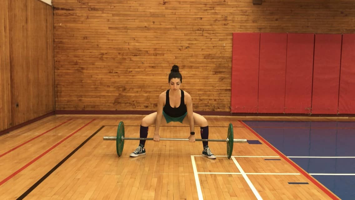 brittany at the bottom of the sumo deadlift, bent over with a flat back, grabbing the barbell in her outstretched hands and in contact with her shins and her chest up