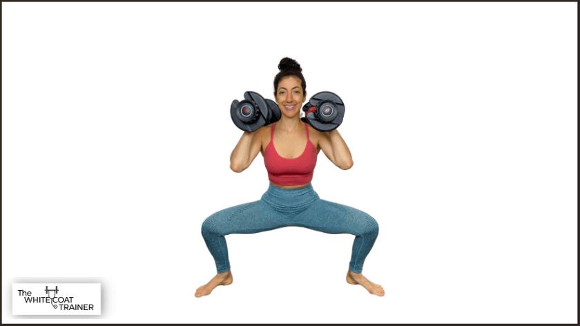 brittany performing a squat with a wide stance and feet pointed out and holding dumbbells on her shoulders
