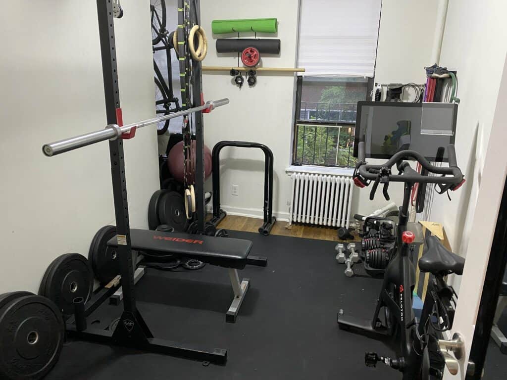 Picture of home gym showing rack, barbell, plates, bench, peloton and more